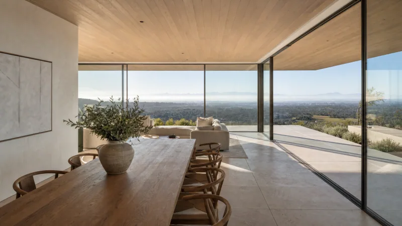 Concept visualisation, not an existing residence: a long oak dining table with woven chairs under an oak ceiling, beside a frameless glass corner that opens onto a stone terrace and a wide valley view.